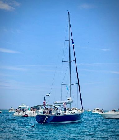 Blue-hulled sailboat with tall mast anchored among several yachts in clear turquoise coastal waters, groups of people on deck enjoying a sunny summer boating day