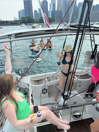 Sailboat summer party on Lake Michigan with Chicago skyline in the background, friends in swimsuits, paddleboards and colorful inflatables, American flag flying