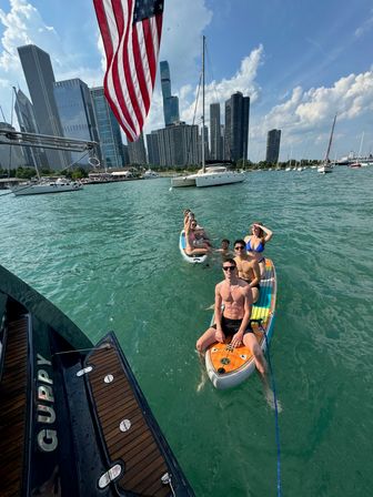 Group of friends lounging on a colorful paddleboard and floating board in turquoise water with sailboats nearby, an American flag overhead and Chicago skyscrapers under a sunny blue sky.