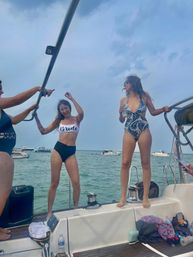 Boat party on a sailboat — friends celebrating a bachelorette, woman in a 'Bride' top dancing and another in a patterned swimsuit with a drink, boats on coastal waters under a partly cloudy sky.