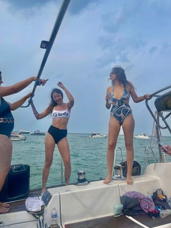 Boat party on a sailboat — friends celebrating a bachelorette, woman in a 'Bride' top dancing and another in a patterned swimsuit with a drink, boats on coastal waters under a partly cloudy sky.