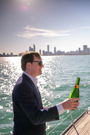 Man in blazer and sunglasses holding a champagne bottle on a sailboat, sparkling water and a city skyline in the background.
