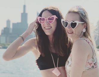 Two smiling women in heart-shaped sunglasses and summer swimwear posing on a boat with a waterfront city skyline in the background.