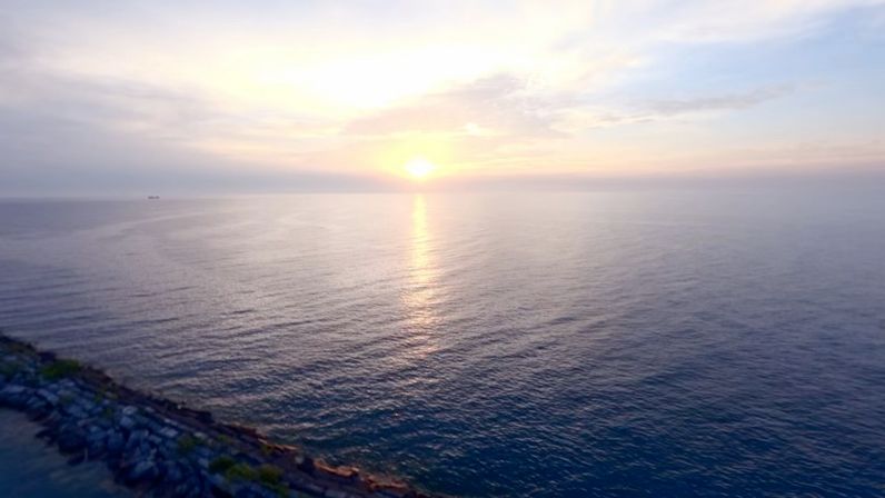 Golden sunrise glinting over a calm open ocean, sunlight reflected on rippled water with a rocky shoreline in the foreground