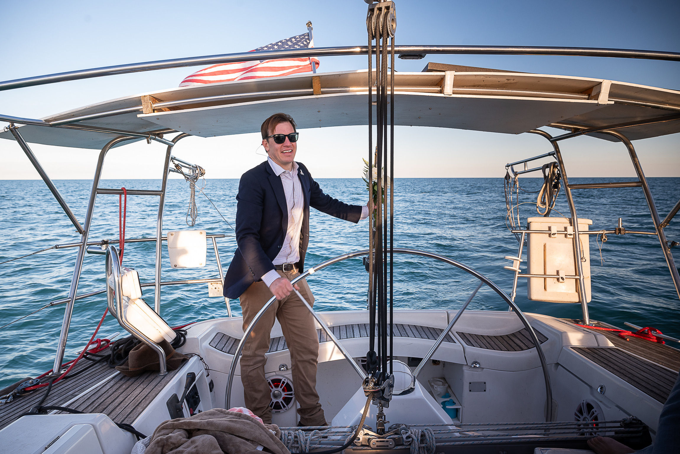 Sunlit person in a blazer and sunglasses steering a sailboat helm under an American flag, cruising on calm blue open sea from the yacht's cockpit.