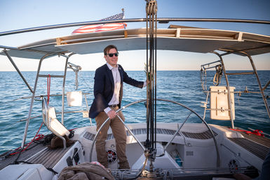 Sunlit person in a blazer and sunglasses steering a sailboat helm under an American flag, cruising on calm blue open sea from the yacht's cockpit.
