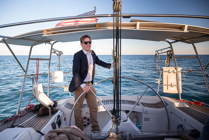 Sunlit person in a blazer and sunglasses steering a sailboat helm under an American flag, cruising on calm blue open sea from the yacht's cockpit.