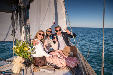 Three smiling adults in sunglasses sitting on a sailboat deck in calm blue ocean under a clear sky, flashing peace signs beside a wrapped bouquet of yellow roses and a cozy blanket.