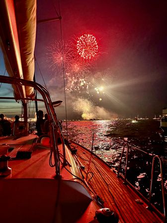 Sailboat deck at night watching red fireworks burst over a busy harbor, bright bursts reflecting on rippling water and silhouetted passengers
