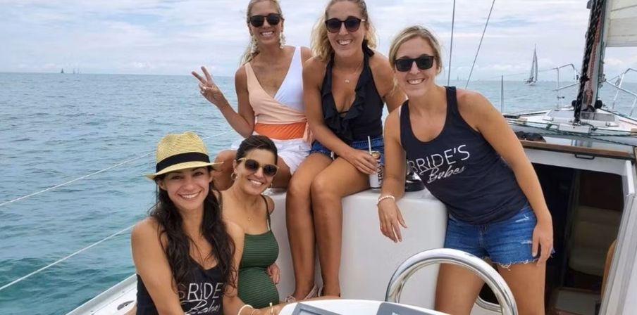Five women in sunglasses and summer outfits smiling on a sailboat deck in calm blue coastal waters with other sailboats on the horizon — bride-themed bachelorette outing.