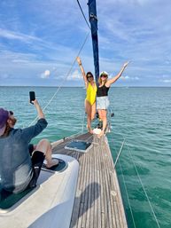 Two friends posing on the bow of a sailboat over turquoise ocean waters and blue sky — one in a yellow swimsuit, the other in a black top, denim skirt and captain’s hat, while a person photographs them from the deck.