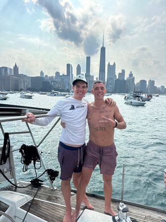 Two friends posing on a sailboat deck with the Chicago skyline and multiple boats on Lake Michigan on a hazy summer day.