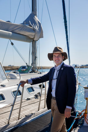 Smiling man in a wide-brimmed hat and navy blazer standing on a sailboat at a sunny marina, holding the boat's railing with blue sky and calm harbor water in the background.