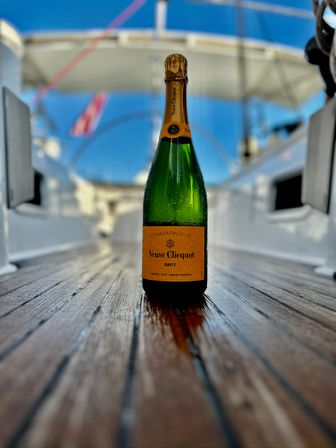 Chilled champagne bottle with an orange label standing on a teak yacht deck, sunlit blue sky and blurred nautical flags in the background
