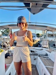 Smiling woman on a sailboat deck at a sunny marina, wearing a gray cap and light shorts, giving two thumbs up with other boats and docks in the background.