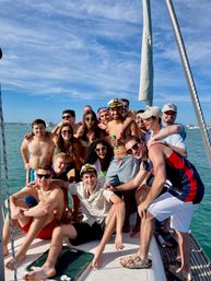 Group of smiling young adults in swimwear and captain hats crowded on a sailboat deck on a sunny day with turquoise coastal waters and other boats in the background