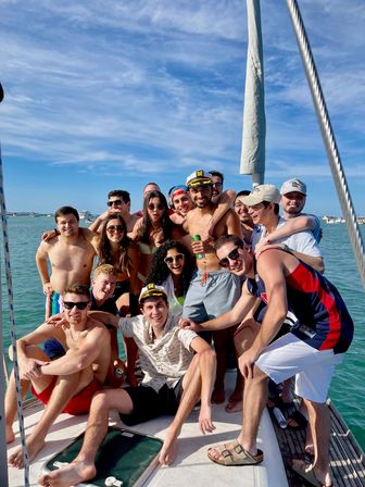 Group of smiling young adults in swimwear and captain hats crowded on a sailboat deck on a sunny day with turquoise coastal waters and other boats in the background