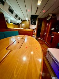 Cozy sailboat cabin interior — glossy wooden dining table and teak floor, green cushioned seating, compact galley with sink and portholes, storage and cooler in a warm yacht salon.