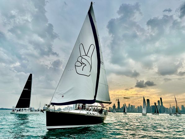 Sailboat with a large peace-sign sail gliding on teal water at sunset with the Chicago skyline and other sailboats under a dramatic cloudy sky