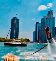 Flyboarder propelled above turquoise water beside a sailboat with a rainbow flag, with downtown Chicago skyline and high-rise buildings on a sunny lakefront day.