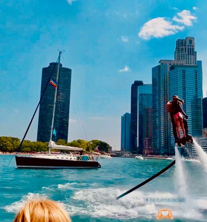 Flyboarder propelled above turquoise water beside a sailboat with a rainbow flag, with downtown Chicago skyline and high-rise buildings on a sunny lakefront day.