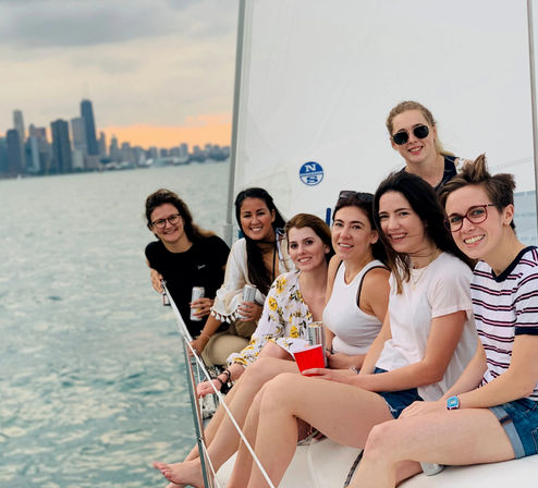 Smiling group of women relaxing on a sailboat with the Chicago skyline over Lake Michigan at sunset, holding drinks.