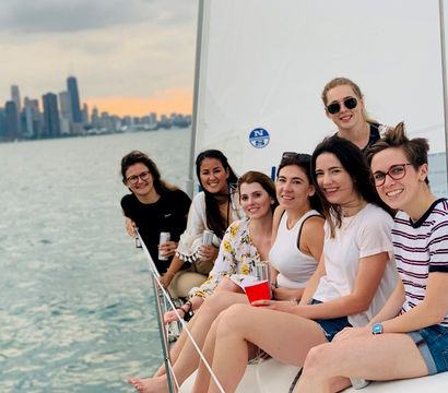 Smiling group of women relaxing on a sailboat with the Chicago skyline over Lake Michigan at sunset, holding drinks.