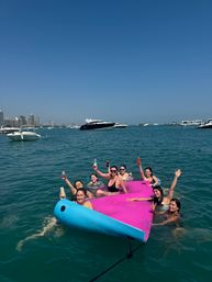 Group of friends lounging and cheering on a bright pink-and-blue inflatable in turquoise harbor waters, holding drinks with yachts and a distant urban skyline under a clear blue sky