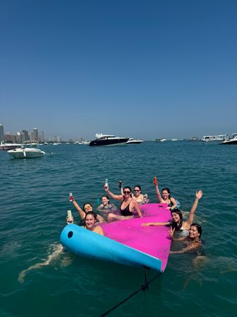 Group of friends lounging and cheering on a bright pink-and-blue inflatable in turquoise harbor waters, holding drinks with yachts and a distant urban skyline under a clear blue sky