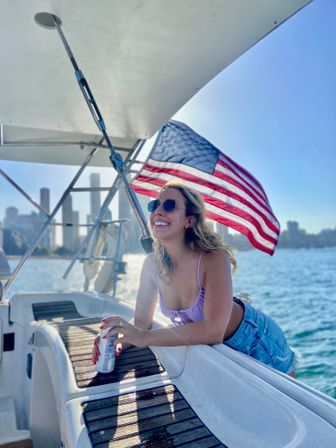 Smiling woman in sunglasses leaning on a sailboat rail with a drink, American flag waving and a city skyline over blue water on a sunny day