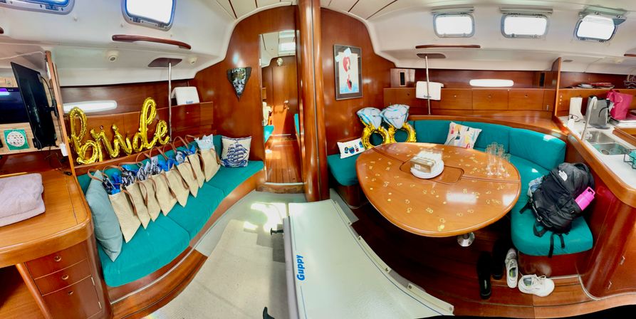 Sailboat interior salon with teal curved seating and wooden table decorated for a bachelorette: gold "bride" balloons, inflatable ring balloons, gift bags, scattered confetti, backpacks and sneakers.