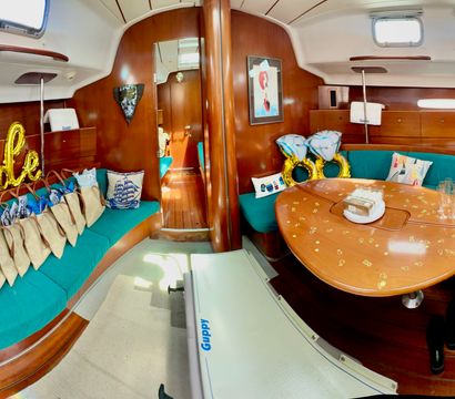 Sailboat interior salon with teal curved seating and wooden table decorated for a bachelorette: gold "bride" balloons, inflatable ring balloons, gift bags, scattered confetti, backpacks and sneakers.