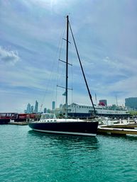 Sleek black sailboat moored at a busy city marina, turquoise water and skyline under a bright, partly cloudy sky.