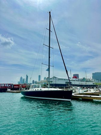 Sleek black sailboat moored at a busy city marina, turquoise water and skyline under a bright, partly cloudy sky.