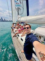 Selfie on a sailboat with a smiling group in swimsuits on deck, Chicago flag and skyline visible over turquoise Lake Michigan waters.