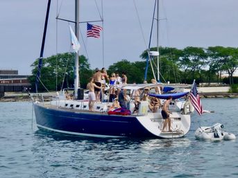Group of friends partying on a navy-hulled sailboat at an urban lakefront harbor, American flag flying, small inflatable dinghy alongside, people in swimsuits enjoying a sunny summer day