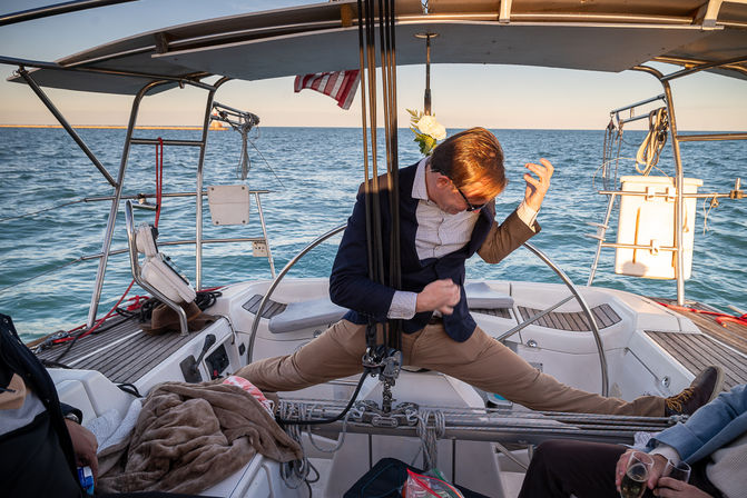 Playful person in a blazer stretching legs across a sailboat cockpit while clutching the helm, calm ocean and an American flag at sunset.