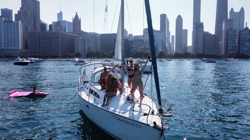 Friends in swimsuits enjoying a white sailboat on blue water with the Chicago skyline behind them on a sunny day.