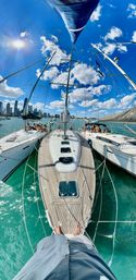 View from a sailboat bow over teak deck into turquoise harbor, three yachts tied side-by-side, sun glinting on water, people relaxing on neighboring boats and a distant city skyline under a bright blue sky with fluffy clouds.