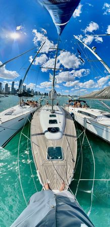 View from a sailboat bow over teak deck into turquoise harbor, three yachts tied side-by-side, sun glinting on water, people relaxing on neighboring boats and a distant city skyline under a bright blue sky with fluffy clouds.