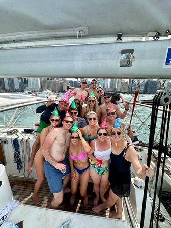 Group of friends in swimsuits wearing colorful birthday party hats and holding drinks, posing and smiling on a sailboat deck with a busy urban waterfront skyline and marina in the background on a sunny summer day.