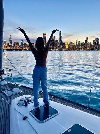 Silhouette of a person standing on a sailboat deck with arms raised, Chicago skyline glowing at dusk across Lake Michigan