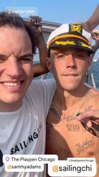 Two smiling people on a sunny Chicago boat selfie — one wearing a captain’s hat and chain, the other in a white sailing shirt, with blue water and sky behind them.