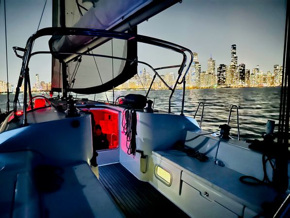 Night sailboat cockpit with red-lit cabin, deck rigging and seating in the foreground and an illuminated city skyline reflected on the water in the background.