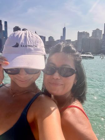 Two women in sunglasses smiling on a boat on Lake Michigan, turquoise water, moored yachts and the Chicago skyline of skyscrapers under a blue sky.