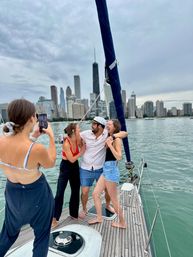 Friends laughing on a sailboat deck on Lake Michigan—one wearing a captain’s hat—while a fourth snaps a photo, with the Chicago skyline and cloudy sky in the background.