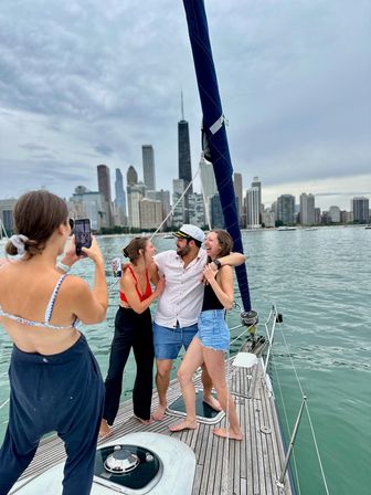 Friends laughing on a sailboat deck on Lake Michigan—one wearing a captain’s hat—while a fourth snaps a photo, with the Chicago skyline and cloudy sky in the background.