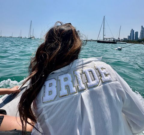 Person wearing a white jacket with 'BRIDE' on the back enjoying a sunny boat ride on an urban harbor with sailboats and a city skyline in the distance