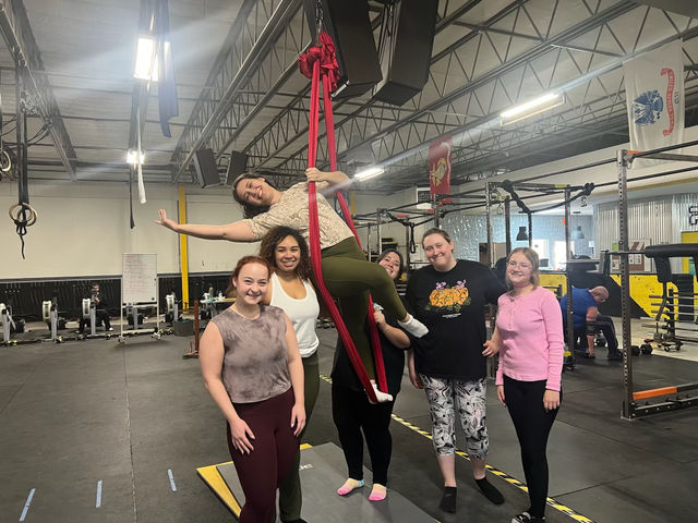 Group of six smiling women in a warehouse-style gym posing for an aerial silks class — one woman suspended in red silks while others stand on rubber flooring near weight racks and rowing machines.