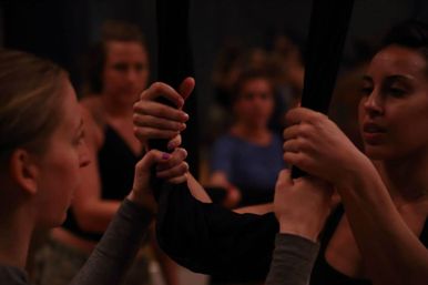 Close-up of women's hands gripping black aerial silks during an indoor aerial yoga class, blurred classmates in the background
